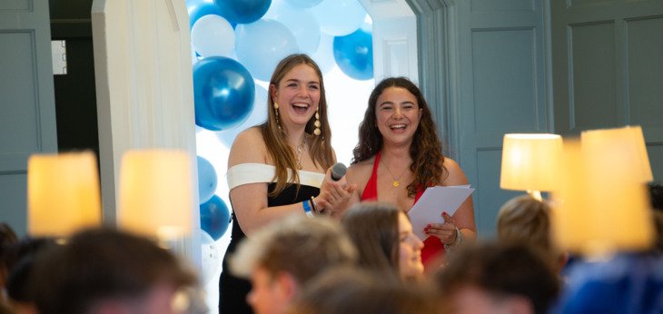  Lower Sixth pupils in black tie holding a microphone to give a speech at the UNICEF Charity Ball in the Dining Room 