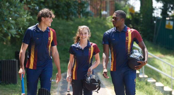  Cricketers in their kit holding cricket equipment walking along the Home Ground 